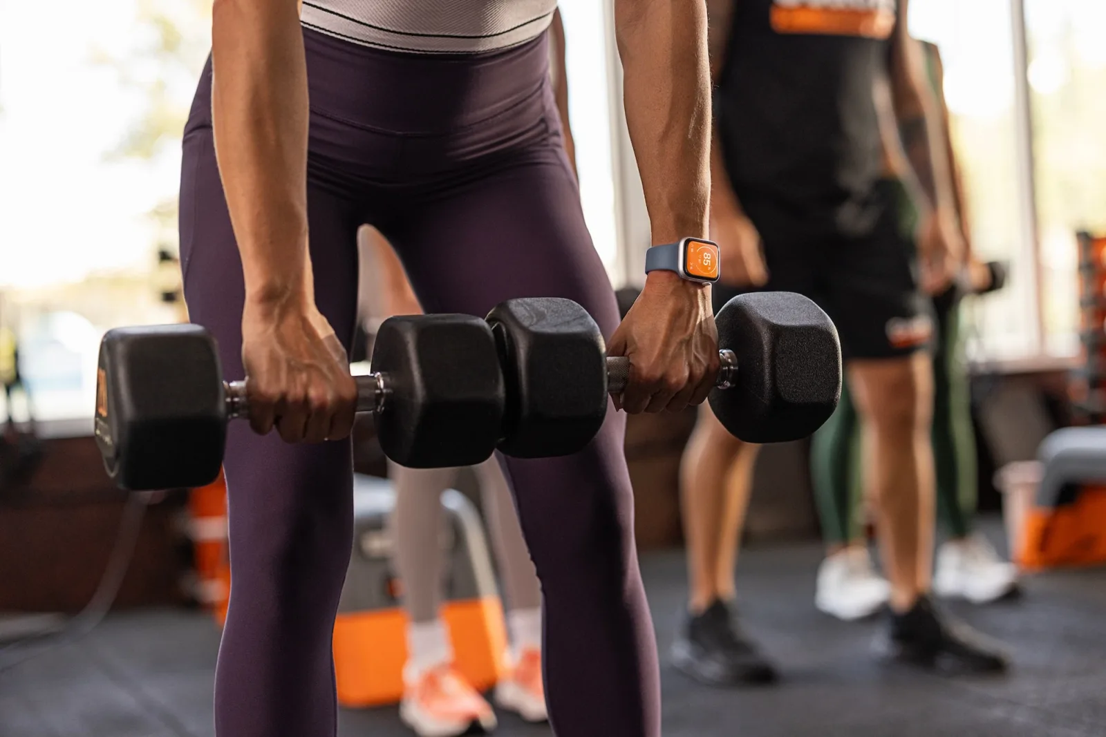 An Orangetheory member lifting dumbbells in class, wearing an Apple Watch that shows the orange Orangetheory workout screen